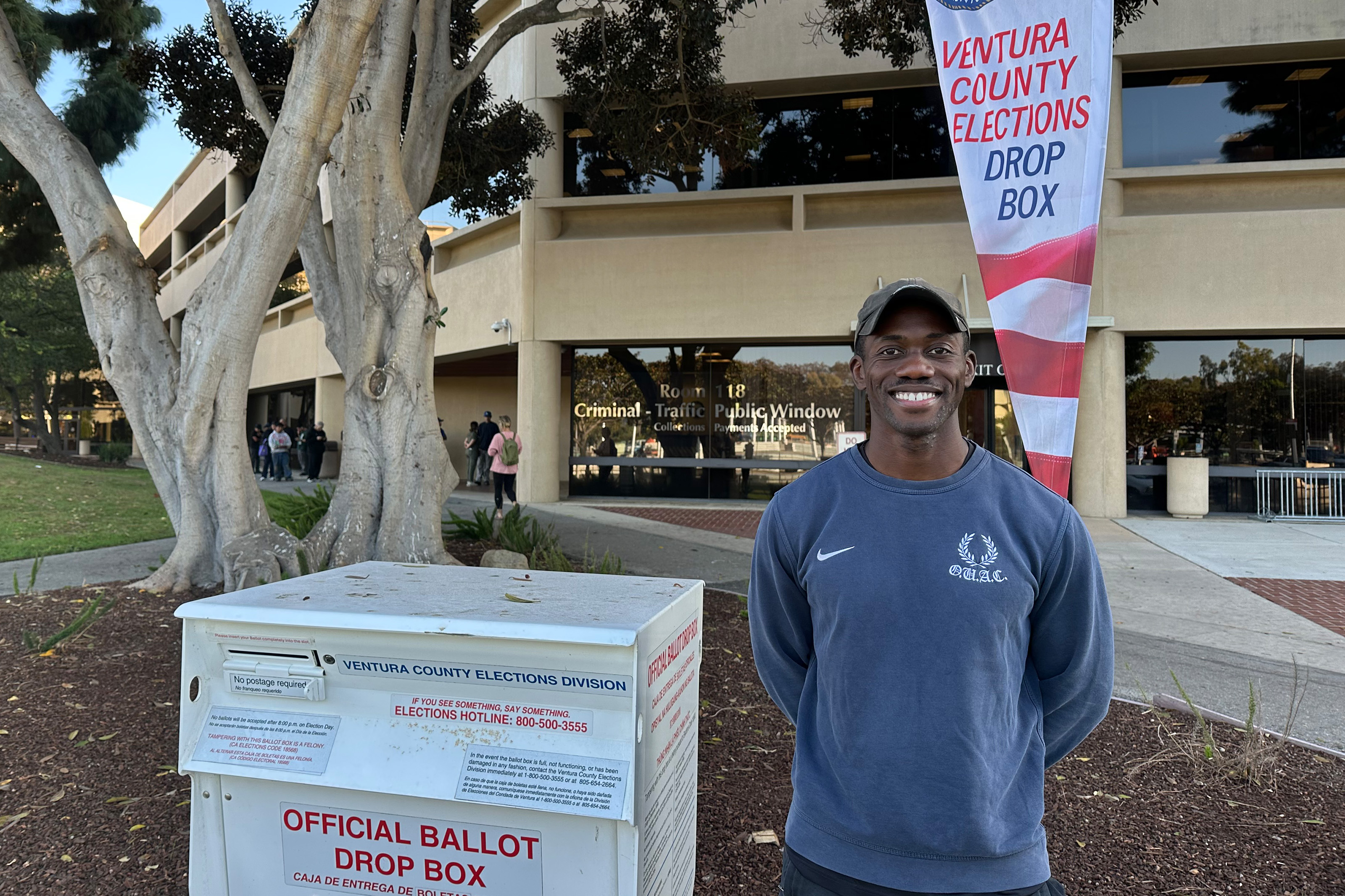 A photo of a man smiling beside a ballot box.