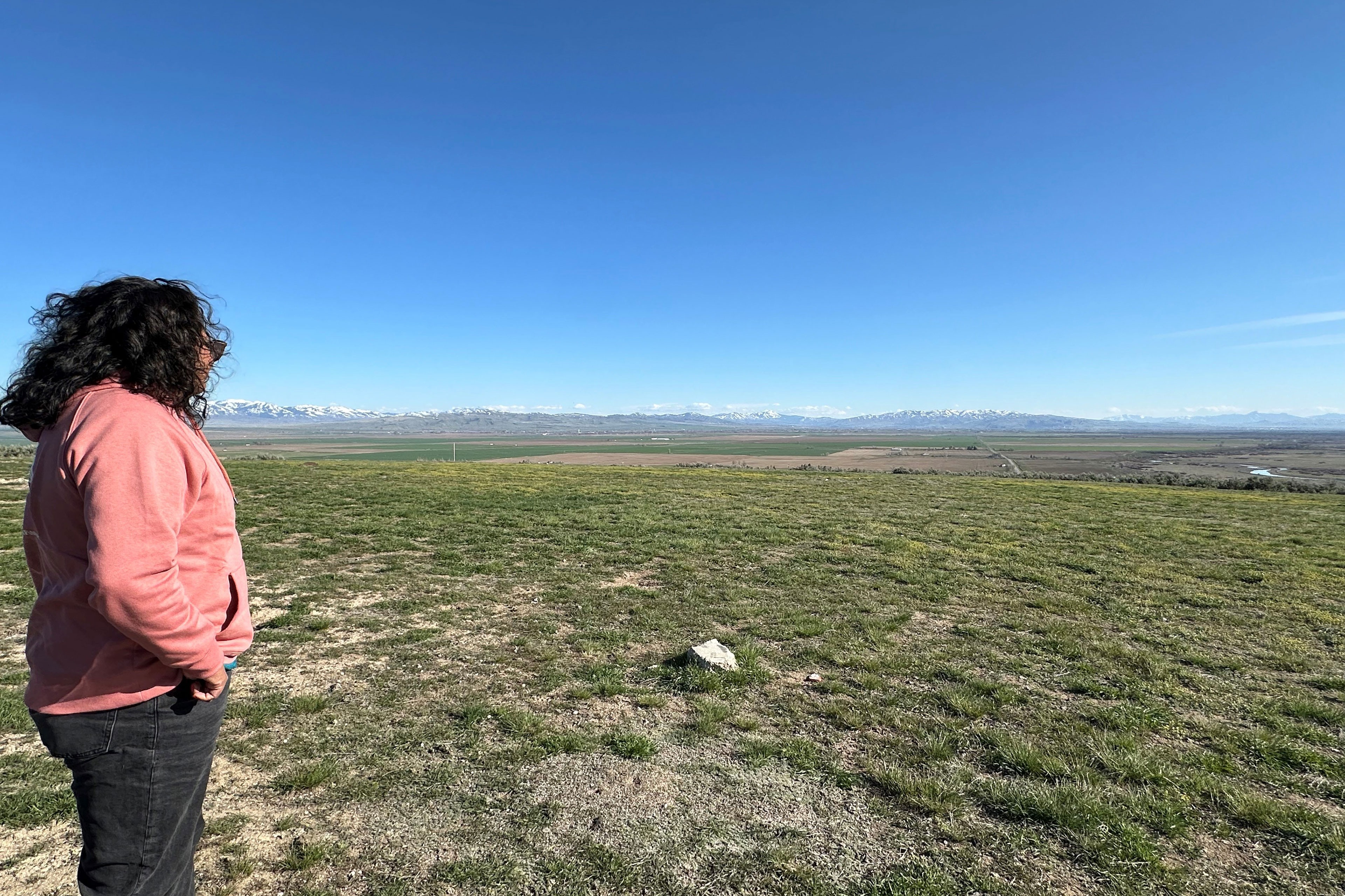 A woman in a pink sweatshirt and black jeans looks out over broad expanse of grassy land. Mountains can be seen in the distance, just above the horizon.