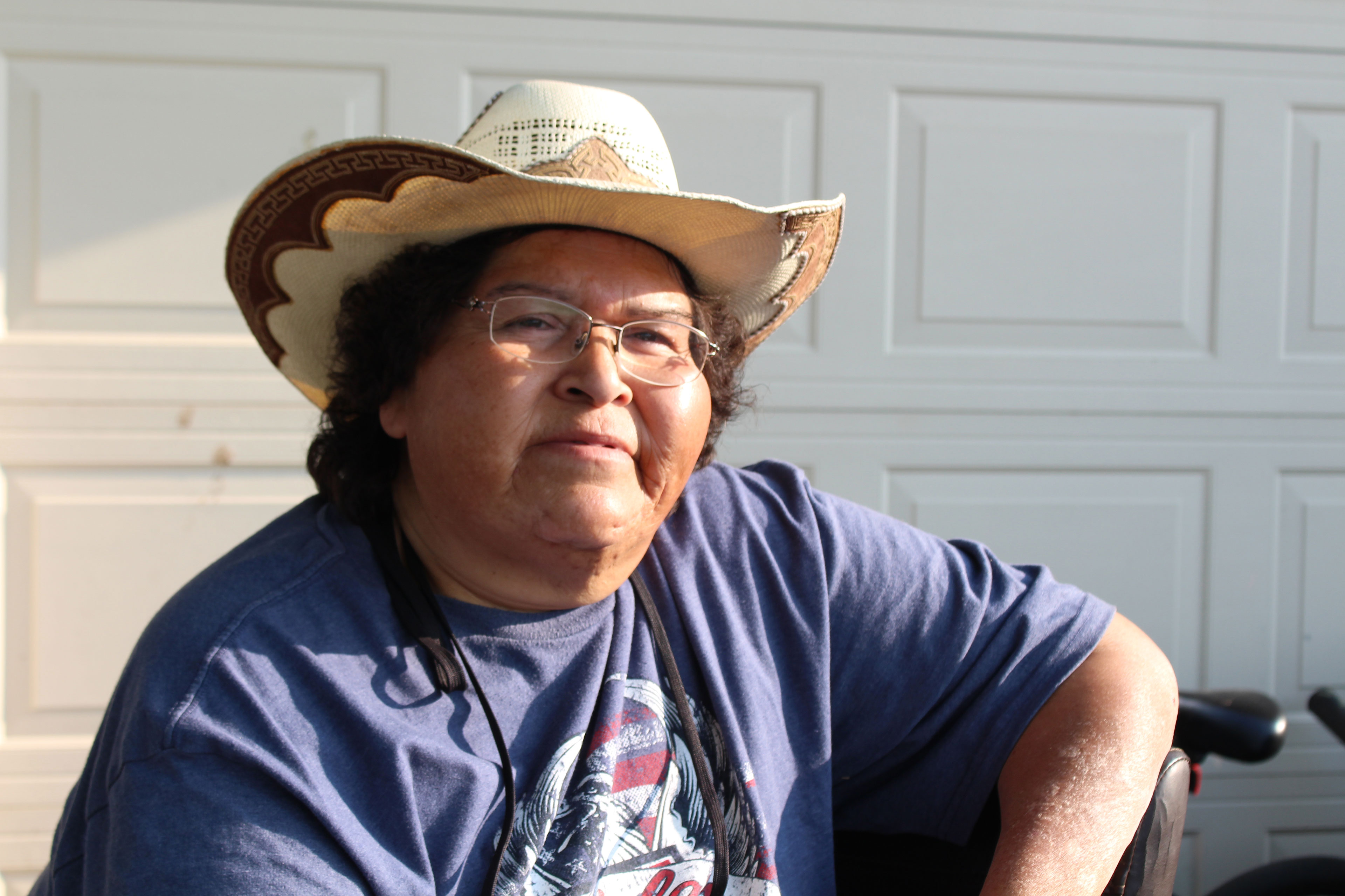 A woman in a brimmed hat and t-shirt sits outside a garage door and looks at the camera.